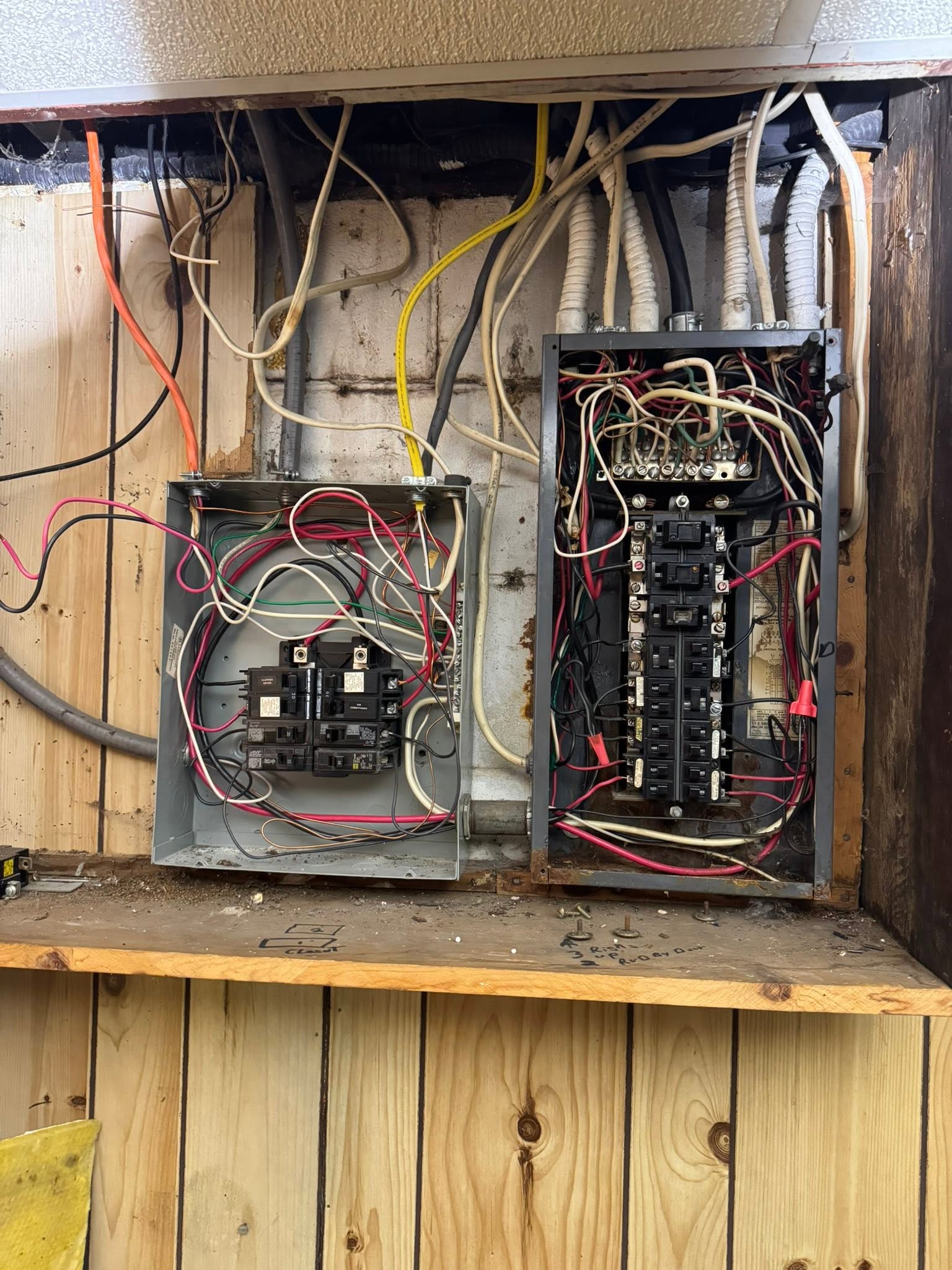 Two open electrical panels with exposed wiring mounted on a wood-paneled wall, showing various cables, circuit breakers, and electrical components.