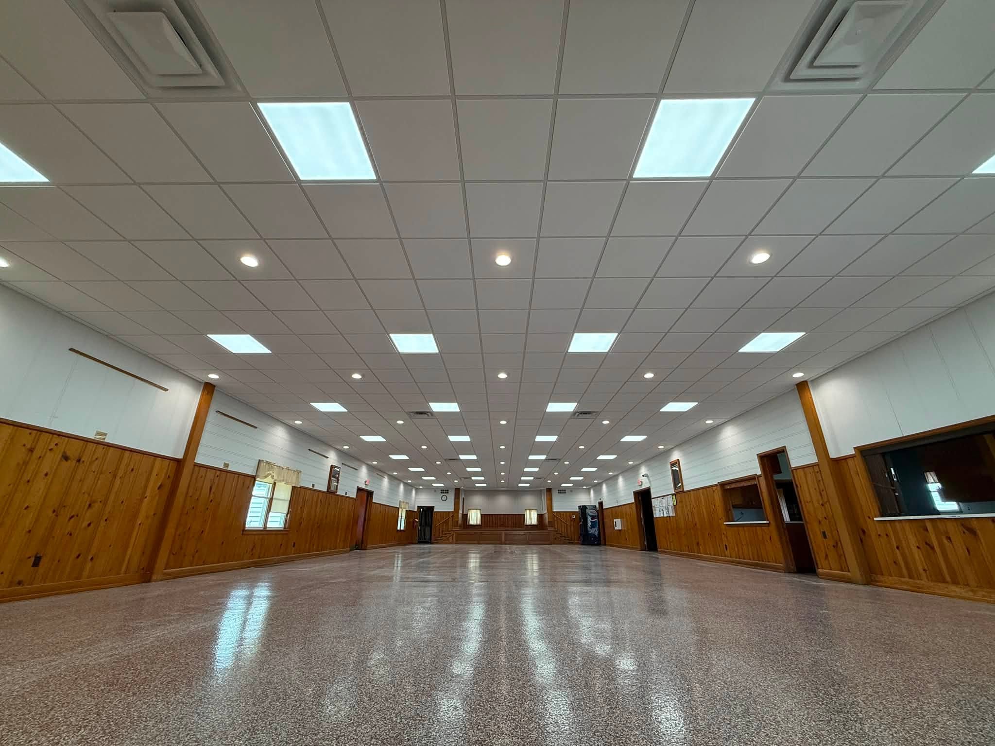 Large, empty hall with wood-paneled walls, white drop ceiling with bright lights, terrazzo floor, and service windows along the sides.