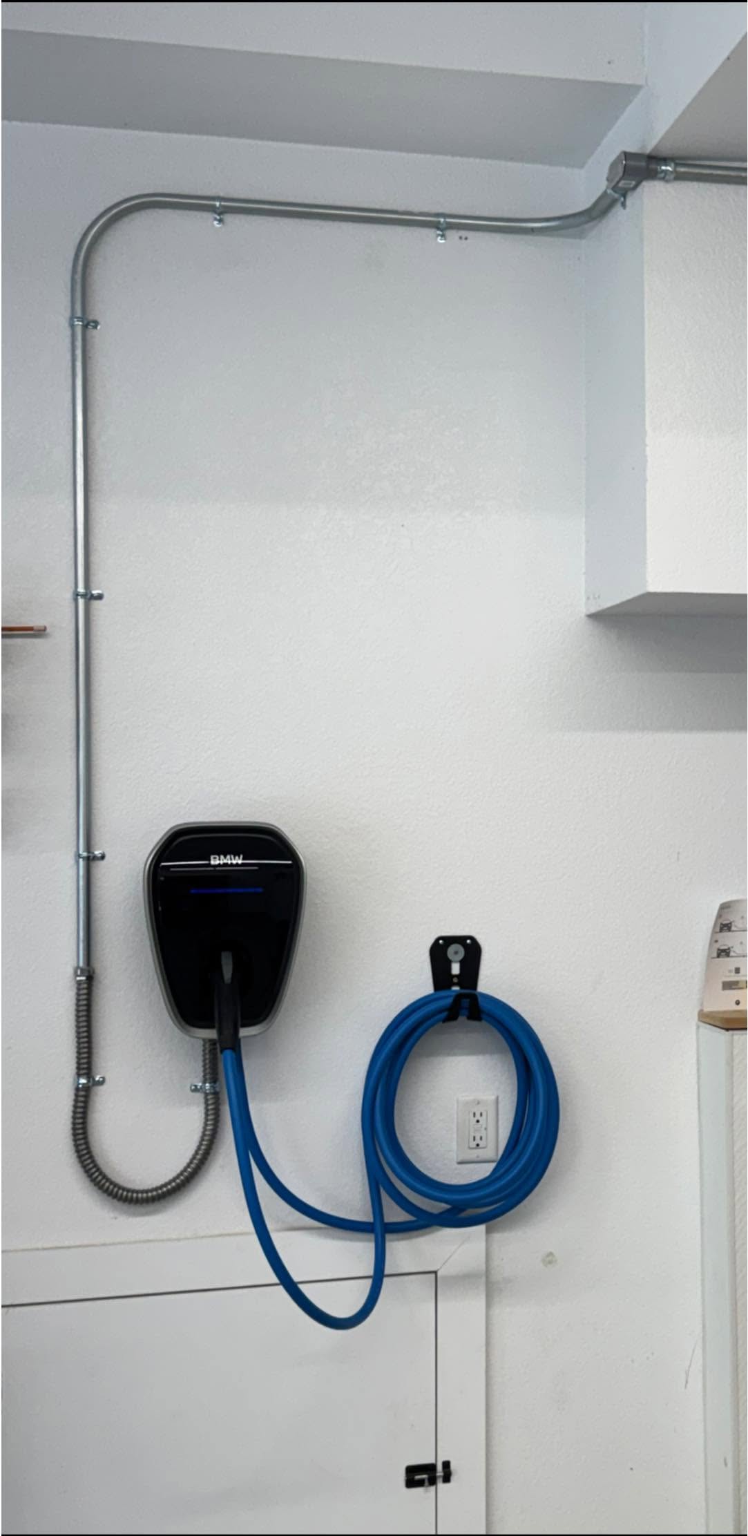 BMW electric vehicle wall charger with a blue charging cable coiled on a holder, mounted on a white wall with visible conduit and an electrical outlet below.