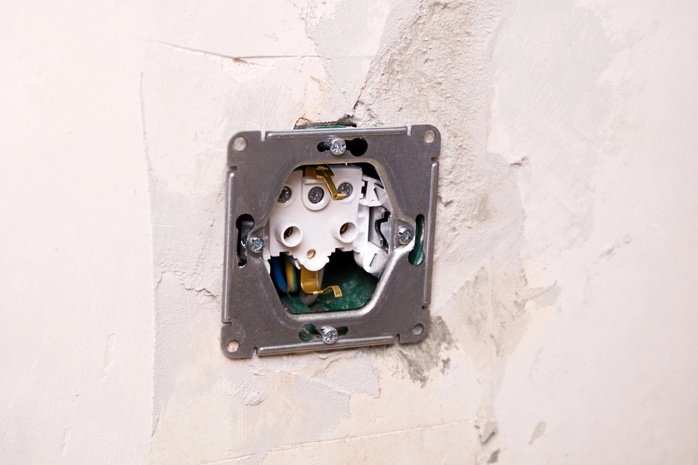 An electrical wall socket without a cover plate is installed in a plastered wall, showing exposed wiring and mounting screws.