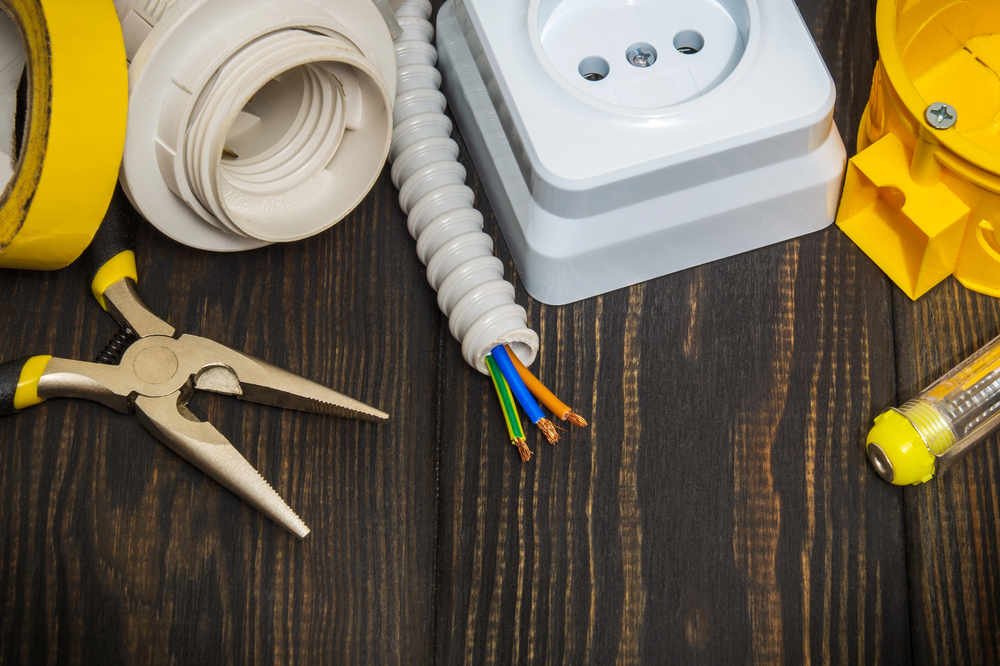 Electrical tools and supplies, including pliers, insulated wires, a wall socket, a yellow box, and a screwdriver, are arranged on a dark wooden surface.