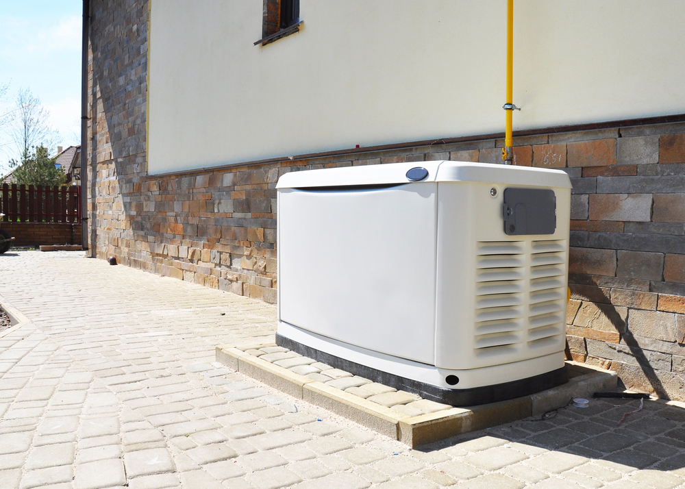 A standby generator is installed on a concrete pad next to a house with stone siding and a paved walkway.