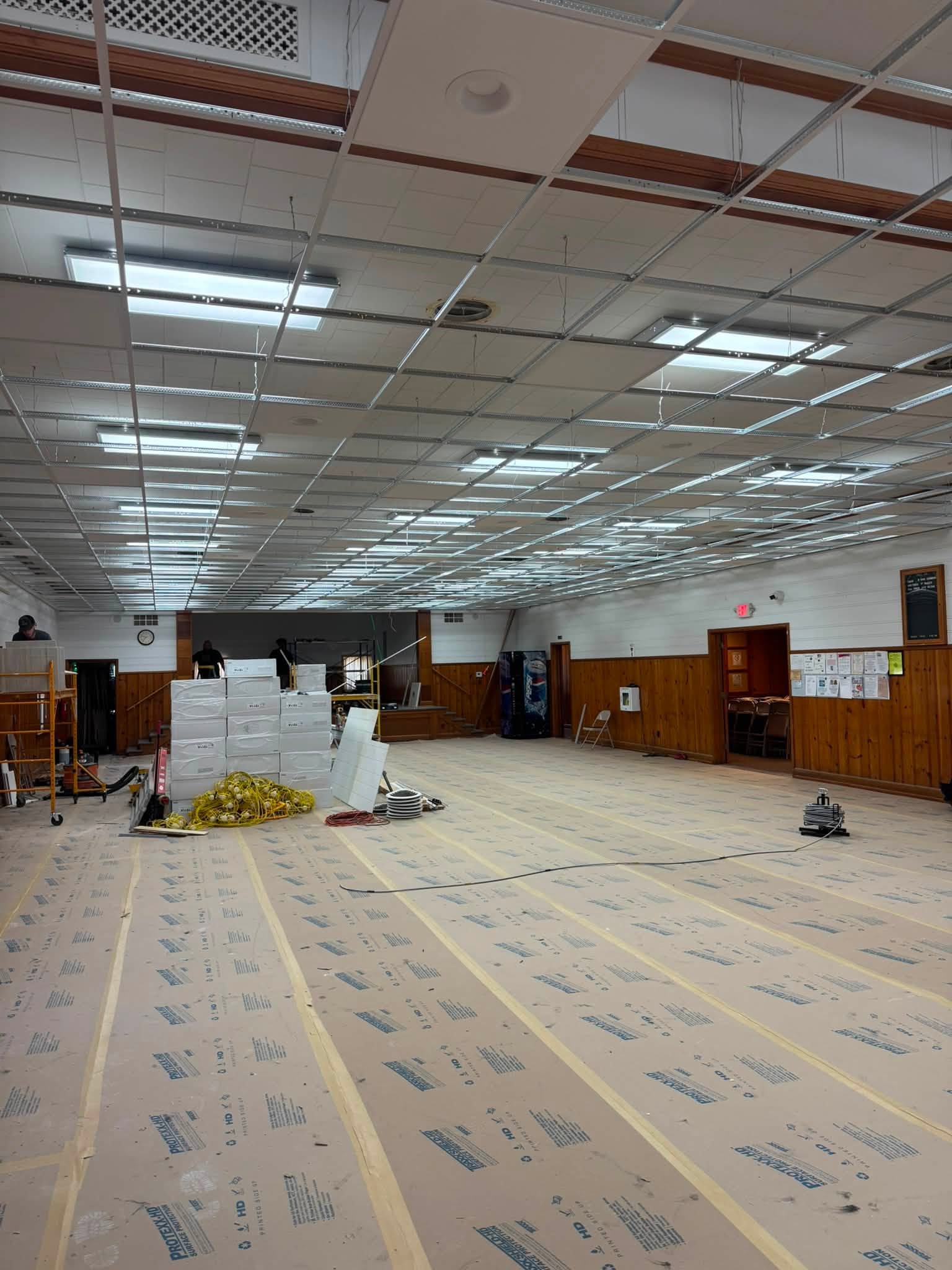 Large room under renovation with ceiling tiles removed, lights exposed, boxes stacked, and protective covering on the floor. Wooden paneling lines the walls, and ladders and tools are visible.