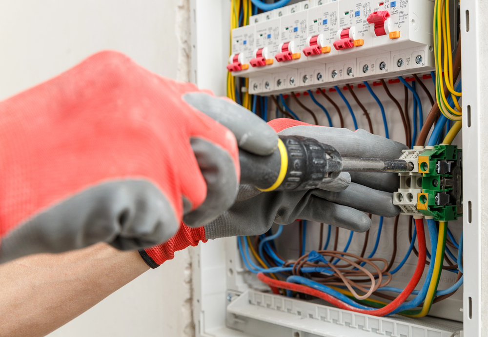 Person wearing red gloves uses a screwdriver to work on an electrical panel with colored wires and circuit breakers.