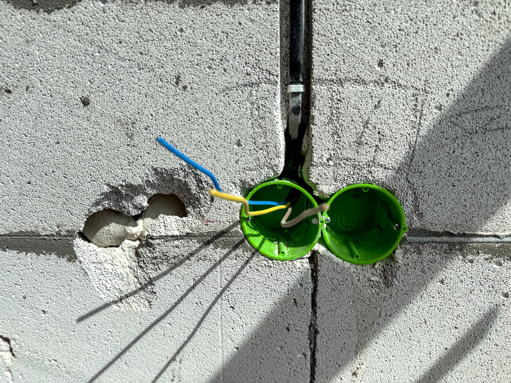 Two green electrical boxes are set into a concrete wall with three exposed wires—blue, yellow, and white—extending from them. A hole is visible in the wall on the left side.